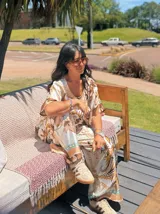 Pantalón palazzo de satén blanco con estampado barroco de hojas tropicales en tonos verde oliva y mostaza, y detalles de cadenas en color dorado y rosado. Presenta un borde inferior con detalles en tonos melocotón y azul claro.