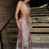 Vestido largo de fiesta con escote halter, espalda descubierta y abertura lateral en la falda. Presenta un top triangular con detalle de abertura en el abdomen y una cadena dorada con dije de concha que rodea la cintura. El estampado es abstracto en tonos grises y marrones.