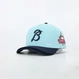 Gorra de béisbol con diseño de doble visera, en color celeste y azul marino. Presenta una letra B bordada en el frente, el texto Desires bordado en el lateral y un parche gráfico con el nombre de la marca y un skyline urbano.