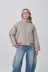 Campera acolchada de diseño capitoné en color beige, con cuello alto, cierre frontal y bolsillos laterales. Presenta un corte holgado con detalles de costuras reforzadas en las mangas y ajuste en la cintura.