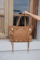 Bolso estilo shopper de gamuza color marrón, con diseño artesanal que incluye flecos laterales y apliques de tachas metálicas circulares. Cuenta con doble asa de hombro y costuras a la vista.