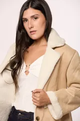 Abrigo tipo gamulán de corte largo, color beige con interior y detalles en corderito sintético color crema. Presenta cierre frontal con botones, cuello solapa y bolsillos cuadrados frontales.