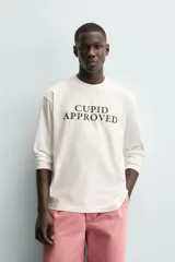 Camiseta de manga larga color blanco roto, corte relajado (relaxed fit) con cuello redondo y texto estampado en negro en el pecho que dice "CUPID APPROVED".