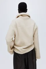 Campera de sherpa suave con cuello elevado, cierre frontal y bolsillos laterales de parche. Presenta un diseño de hombros caídos y corte holgado.