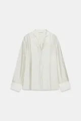 Camisa fluida de mujer color blanco crudo, confeccionada en mezcla de lyocell. Tiene escote en pico con cuello solapa, manga larga con puños anchos y cierre frontal oculto con botones.