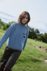 Sweater de punto color marrón con cuello alto, manga larga y calce relajado. Presenta detalles bordados en color mostaza en el cuello, puños, ruedo y bolsillo frontal, además de un aplique en forma de corazón en el bolsillo.