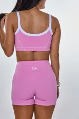 Conjunto deportivo compuesto por un top y un short de tiro alto en color rosa. El top presenta breteles finos blancos, escote redondo y el logo 'bk.' estampado en el centro. El short cuenta con costuras decorativas blancas que resaltan el diseño.