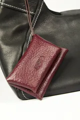 Cartera de cuero vacuno color negro, con costuras a contratono en color blanco y un práctico bolsillo frontal desmontable en color bordó con tiras envolventes. Cuenta con asa para el hombro.