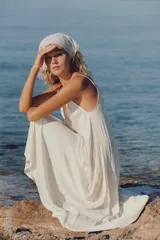 Vestido largo satinado color crudo, con escote en pico adornado con lazada y tirantes finos ajustables. Presenta detalles de frunces y diseño fluido.