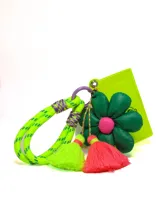 Llavero con cordón trenzado verde neón, detalles en violeta y dijes colgantes: flor verde con centro rosa, borla verde neón y borla fucsia. Incluye tarjetero de plástico rígido color verde neón.