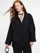 Camisa negra de corte oversize y manga larga con puños abotonados. Presenta un cuello clásico con un lazo fino para anudar en la parte superior y un diseño con bajo asimétrico, siendo más larga en la parte posterior.