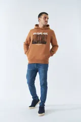 Buzo marrón con capucha y bolsillo canguro. Presenta un estampado frontal con el logo de la marca Mistral y una imagen de un arco arquitectónico. Confeccionado en algodón y poliéster.
