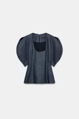 Blusa de Zara confeccionada en mezcla de algodón con acabado denim. Presenta escote redondo, mangas cortas abullonadas con volumen y costuras con pespuntes a contraste. Cierre posterior con cremallera oculta.