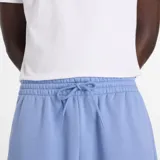Pantalón de jogging New Balance color azul claro, confeccionado en tejido polar suave. Presenta un corte relajado, cintura elástica con cordón ajustable y puños elásticos en los tobillos. Incluye el logotipo de la marca bordado en la pierna izquierda.