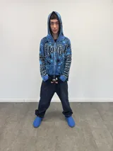 Buzo tipo hoodie tejido de punto grueso con cierre frontal completo hasta la capucha. Presenta un diseño gráfico abstracto en tonos azules y negros, con tipografía estilo gótico en el frente y las mangas. Cuenta con puños y ruedo acanalados en color negro, y etiqueta de marca en la manga.
