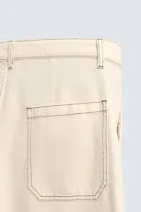 Pantalón de algodón color beige claro, de corte recto (regular fit), con pespuntes en hilo contrastante oscuro. Presenta bolsillos delanteros y bolsillos traseros de parche.