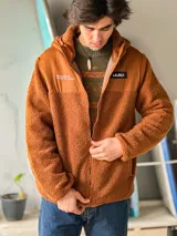Campera de corderito de manga larga y cierre frontal, con diseño en bloques de color azul y negro. Cuenta con capucha y bolsillos laterales, además de detalles estampados con la marca y la frase 'boardbrand made for riders'.