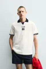 Polo de corte relajado confeccionado en piqué de algodón color blanco. Presenta cuello solapa en color negro con abertura frontal, mangas cortas con ribetes a contraste y estampado conmemorativo USA 94 FIFA World Cup.