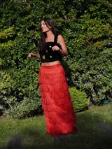 Falda larga de lino color rojo, con aberturas laterales y múltiples niveles de volados formados por flecos de rafia. Cuenta con cintura elástica en la parte posterior.