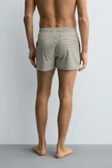 Bañador corto de hombre con cintura elástica ajustable mediante cordón blanco. Presenta un estampado geométrico repetitivo en tonos oscuros (negro/marrón oscuro) y blanco/beige. Incluye bolsillos laterales y un bolsillo trasero de vivo.