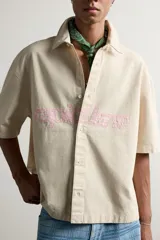 Sobrecamisa boxy fit de algodón color beige, de manga corta, con cuello solapa, cierre frontal de botones y aplicación de texto en parches rosas a contraste en el delantero.
