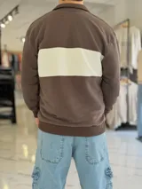 Hoodie de corte clásico con medio cierre y cuello alto, en color marrón con una franja horizontal blanca en el pecho que incluye el logo estampado en negro. Presenta además un pequeño bordado en el hombro izquierdo y puños y cintura elastizados de rib.