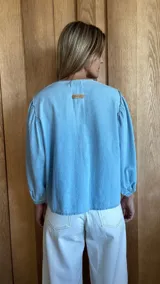 Camisa de jean celeste con cuello redondo, mangas abullonadas y puños elásticos. Cierre frontal con botones.