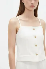 Top blanco de poliéster con tirantes finos, escote recto, entallado con pinzas y cierre frontal con botones dorados con textura.