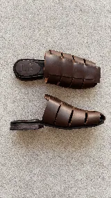Zuecos de cuero marrón con diseño de tiras anchas entrelazadas estilo romano, punta cuadrada y plantilla plana negra con logo grabado de la marca.