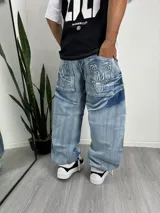 Pantalón de jean estilo baggy, de corte holgado y tiro medio, con lavado claro y efecto desgastado. Presenta bolsillos traseros amplios con bordados decorativos en estilo tribal.