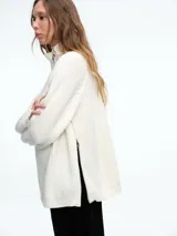 Sweater de tejido de punto acanalado en color blanco, con cuello alto y cierre metálico diagonal en el hombro. Presenta mangas largas con diseño raglán y corte holgado.