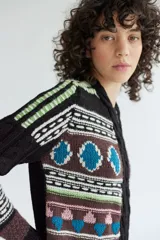 Campera de punto jacquard con lana merino, diseño con cierre frontal, cuello redondo y detalles de trenzas en los laterales. Presenta un patrón geométrico multicolor con tonos marrones, negros, azules y verdes.