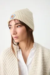 Gorro tipo beanie de punto acanalado en color crudo, con parche circular bordado que presenta un sol estilizado.