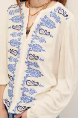 Camisa de color crudo con bordados en tonos azules de diseño paisley en el frente y las mangas. Presenta escote en V con abotonadura parcial y mangas largas.