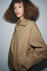 Chaqueta tipo bomber de color beige, con cuello solapa y aplicación extraíble de pelo sintético. Presenta cierre frontal con botones a presión ocultos, bolsillos delanteros y bajo con elástico.