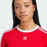 Remera corta roja ajustada al cuerpo, de manga corta, con cuello redondo blanco y las icónicas tres líneas blancas de Adidas en las mangas. Cuenta con el logo del trifolio bordado en el pecho.