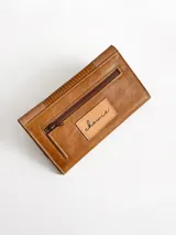 Billetera de cuero color camel con solapa frontal decorada con costuras en forma de cruz y textura grabada. Cuenta con cierre de broche y diseño rectangular.