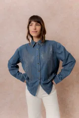Camisa de denim liviano con calce relajado, cuello camisero clásico y cierre frontal con botones. Presenta una destacada aplicación de strass en el frente formando un diseño de alas.