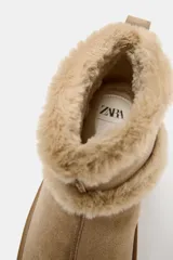 Botín plano de gamuza color beige, con interior y bordes forrados en pelo sintético. Presenta suela de plataforma engomada y tirador trasero.