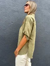 Camisa de gamuza sintética liviana en color verde oliva, con cuello clásico, cierre frontal de botones y mangas cortas con dobladillo.