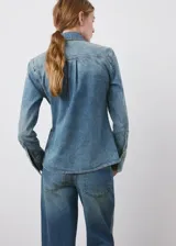 Camisa de denim estilo vaquero, con cuello camisero, manga larga con puños abotonados, cierre frontal con botones y detalle de volantes en el frente.