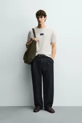 Camiseta de corte relajado, color gris jaspeado, con cuello redondo y manga corta. Presenta un parche rectangular azul marino en el centro del pecho con texto estampado en blanco y una frase adicional debajo.