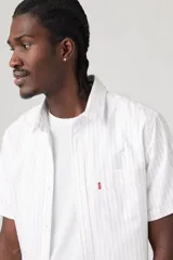 Camisa blanca a rayas verticales finas, de manga corta, con cuello clásico, cierre frontal con botones y un bolsillo en el pecho.