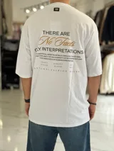 Remera oversize color beige, con mangas cortas y cuello redondo. Presenta un estampado tipográfico en la espalda con la frase "There are no facts, only interpretations" y otros detalles de diseño.