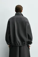 Sudadera de corte relajado color negro, con cuello alto y cierre frontal de cremallera. Presenta un bolsillo de vivo en el pecho con un estampado a contraste y acabados elásticos en puños y ruedo.