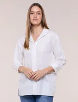 Camisa de silueta holgada y largo extendido, confeccionada en poplín elástico de color blanco. Presenta cuello solapa abierto en V con cartera frontal de abotonadura oculta, hombros caídos y mangas tres cuartos. Cuenta con espalda con canesú, pliegue central y ruedo recto con pequeñas aberturas laterales.