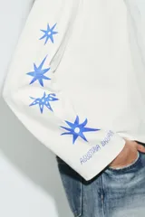 Remera de manga larga y corte oversize, color blanco, con cuello redondo. Presenta un estampado gráfico en color azul en el pecho y a lo largo de la manga derecha.