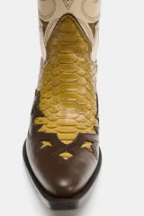 Bota estilo cowboy de caña alta, confeccionada en una combinación de cuero marrón oscuro, beige y texturizado efecto piel de serpiente en tonos mostaza. Presenta bordados decorativos, tacón medio en bloque y puntera perfilada.