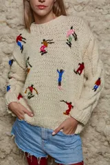 Sweater de lana color crema, tejido a mano con cuello redondo y mangas largas con trenzas. Presenta bordados de figuras esquiando en varios colores.