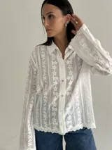 Camisa blanca de manga larga con delicados bordados florales verticales y detalles de encaje. Presenta cuello clásico, cierre frontal con botones y terminaciones festoneadas en los puños y el ruedo.